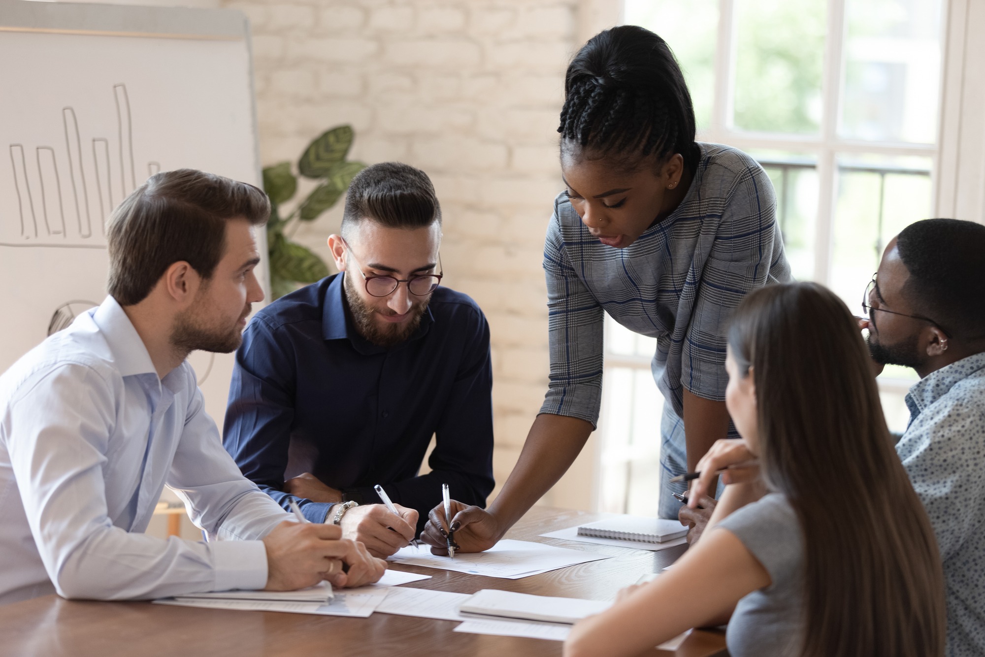 Five colleagues collaborate around a table, focused and engaged. A woman points to documents, directing the discussion.